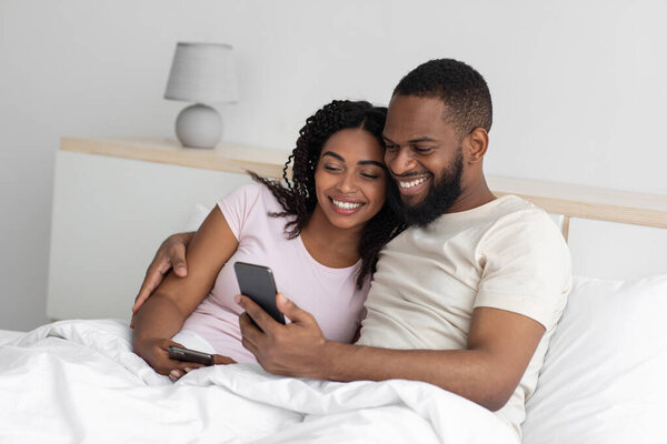 Smiling happy young african american boyfriend and girlfriend hugging, sitting on bed in bedroom interior