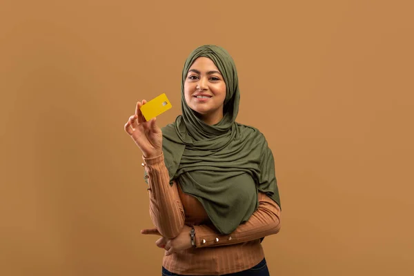 Happy muslim laady in hijab holding credit card advertising bank service, standing over beige background. Easy banking — 图库照片