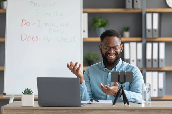 Χαμογελώντας millennial μαύρο δάσκαλο σε γυαλιά ματιά στο φορητό υπολογιστή, κάμερα του τηλεφώνου στο εσωτερικό του γραφείου στο σπίτι — Φωτογραφία Αρχείου