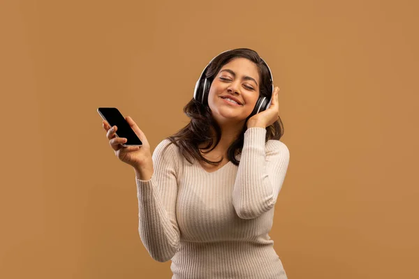 Femme arabe ludique avec écouteurs et smartphone écouter de la musique et profiter d'une playlist cool, fond beige — Photo