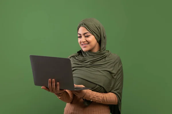 Freelance and online education for muslim women concept. Happy lady in hijab typing on laptop, dark green background — 图库照片