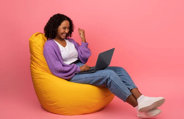 Νίκη στο διαδίκτυο. Ενθουσιασμένη νεαρή μαύρη γυναίκα κάθεται σε καρέκλα φασόλι τσάντα με φορητό υπολογιστή, κάνοντας ΝΑΙ χειρονομία, επιτυγχάνοντας την επιτυχία — Φωτογραφία Αρχείου