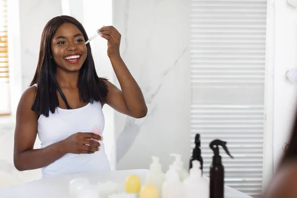 Skin Care. Beautiful Black Female Applying Face Serum Near Mirror In Bathroom — Stock fotografie