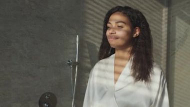 Young positive african american woman wearing bathrobe enjoying sunlight at bathroom, coming to window