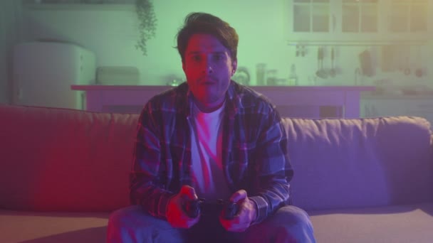 Young millennial guy playing video games with joystick, sitting alone on sofa in neon evening lights, zoom in portrait — Stockvideo