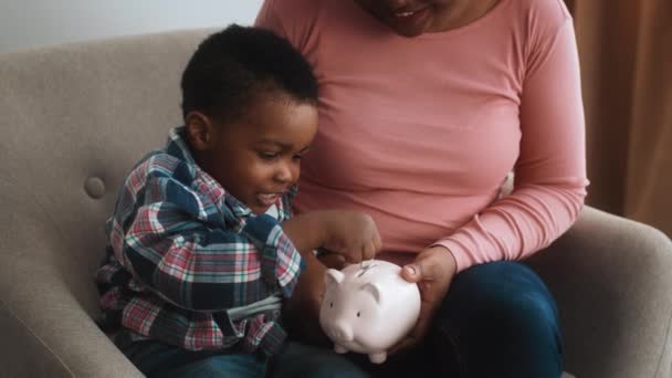 Little black boy putting coins into piggy bank while sitting with mom — Stock video