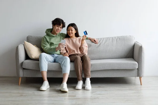 Cheery Asian couple with credit card and smartphone sitting on sofa, ordering goods on internet at home, copy space — Fotografia de Stock