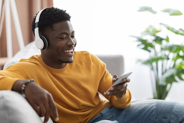 Happy black guy sitting on couch, watching video on smartphone — Zdjęcie stockowe