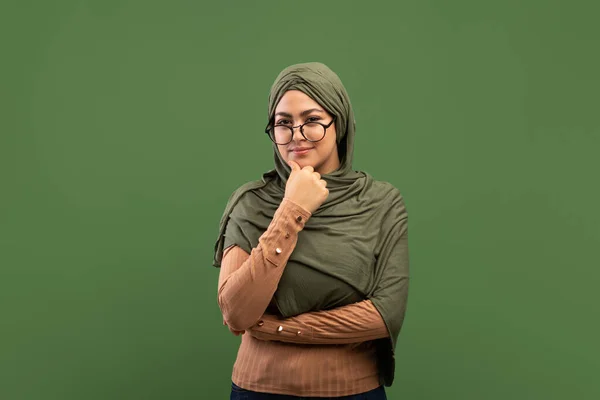 Señora árabe pensativa en gafas tocando la barbilla y pensando en la pregunta, de pie sobre el fondo verde oscuro, espacio de copia — Foto de Stock