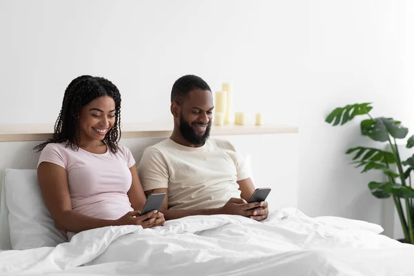 Glad millennial african american guy and lady sitting on bed in bedroom interior, chatting on phones — Fotografia de Stock