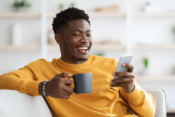Happy african american guy reclining on couch, using smartphone — Foto Stock