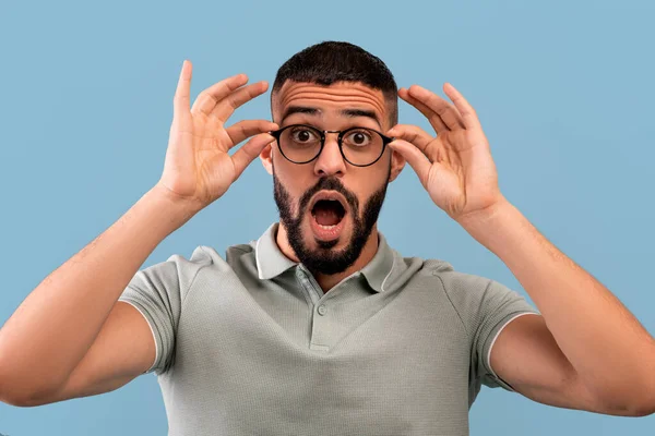 ¡Increíble! Impresionado hombre de Oriente Medio tocando sus gafas y abriendo la boca, mirando a la cámara en el fondo azul — Foto de Stock