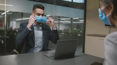 Young man consultant putting on protective mask and greeting woman client, sitting with laptop at office, slow motion