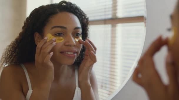 Joven bastante africana americana mujer aplicando anti edema parches de colágeno en la cara, mirando el espejo en el baño — Vídeos de Stock