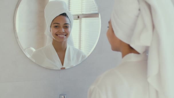 Young african american woman wearing bathrobe and towel on head after shower smiling to camera with mirror reflection — Vídeo de Stock