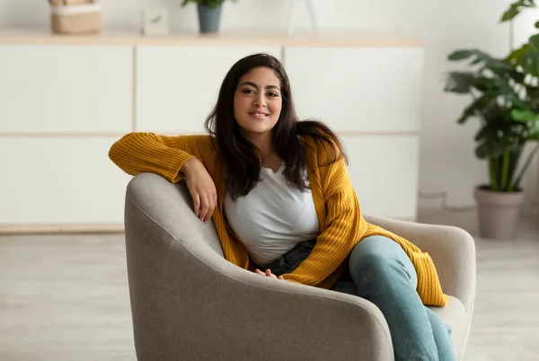 Happy arab lady sitting in comfy armchair and smiling at camera, enjoying lazy morning, having peaceful weekend at home —  Fotos de Stock