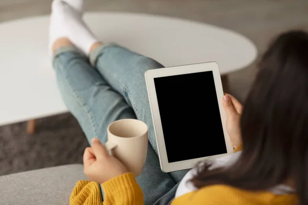 Millennial arab woman using digital tablet with blank screen and holding cup of tea, sitting on sofa at home, mockup — стоковое фото