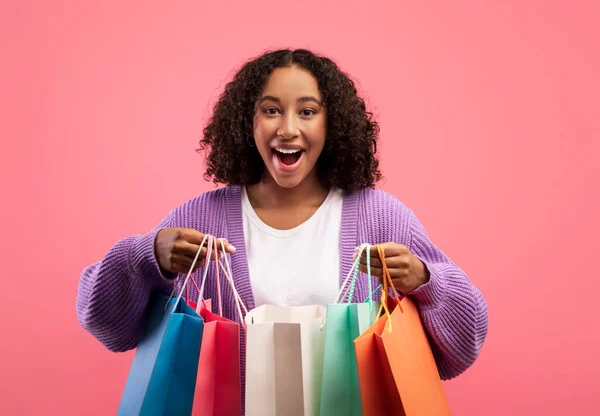 Beautiful millennial black woman looking inside bright shopping bags, happy with her purchases over pink background — 图库照片