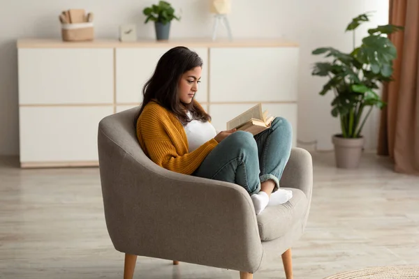 Focused arab lady sitting in armchair, reading book in living room, enjoying lazy morning with interesting story — Foto Stock