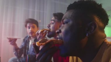 Three multiethnic guys friends eating delivered fresh pizza, watching tv together at home in evening, zoom out portrait