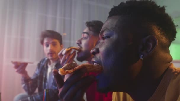 Три багатоетнічні хлопці друзі їдять свіжу піцу, дивлячись телевізор вдома ввечері, збільшують портрет — стокове відео