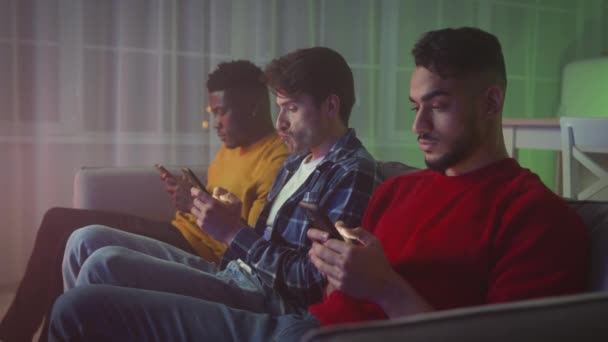 Problems of modern communication. Three multiethnic young men web surfing in social media on phones, ignoring real talk — Wideo stockowe