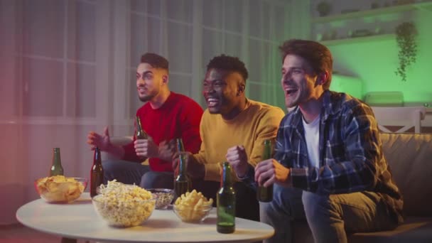 Багатоетнічні хлопці дивляться футбол по телевізору, вітають емоційний футбольний матч з пивом і закусками вдома ввечері — стокове відео