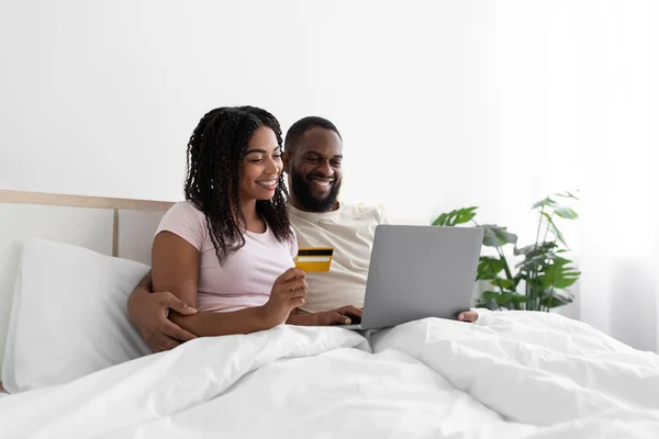 Cheerful millennial african american wife and husband are shopping online, sitting on bed in white bedroom — Stockfoto