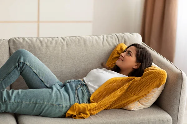 Weekend relaxation. Calm arab lady resting on couch and dreaming, lying on sofa with hands behind head, free space — стоковое фото