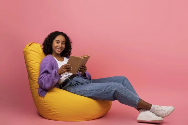 Helg hobbyer. Glada ung svart kvinna med öppen bok sitter i bekväma bönor väska stol på rosa bakgrund — Stockfoto