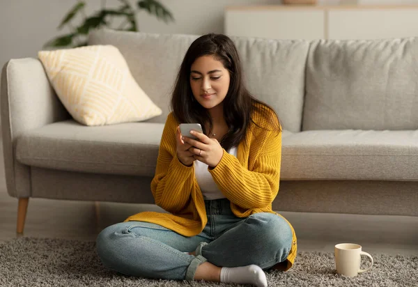 Θετική νεαρή γυναίκα arab διαβάζοντας σε απευθείας σύνδεση πρωινές ειδήσεις στο smartphone και πίνοντας καφέ, κάθεται στο πάτωμα στο σαλόνι — Φωτογραφία Αρχείου