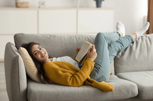 Carefree middle eastern woman resting on couch in living room, reading book while resting at home, smiling at camera — Foto Stock