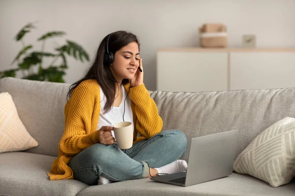 Telecommuting concept. Arab woman sitting on sofa and working online on laptop, wearing headset and holding cup — стоковое фото