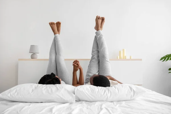Glad happy young african american husband and wife in domestic clothes are lying on bed, holding hands with raised legs — Foto Stock