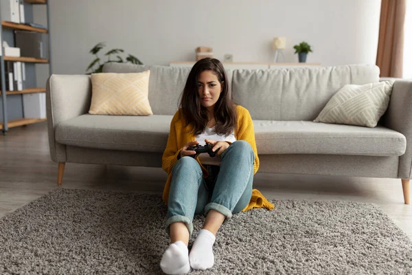 Oyun çubuğuyla oyun oynayan, halının üzerinde oturup bilgisayar oyunlarıyla eğlenen ciddi bir Arap kadın.