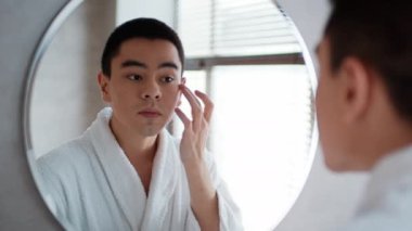 Asian Guy Applying Facial Moisturizer Looking At Mirror In Bathroom