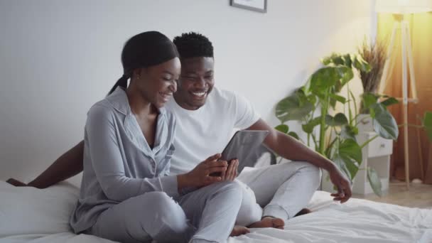 Αστείο περιεχόμενο στο διαδίκτυο. Νεαρός ευτυχισμένος αφροαμερικανός άνδρας και γυναίκα βλέποντας social media σε ψηφιακή ταμπλέτα στο κρεβάτι — Αρχείο Βίντεο