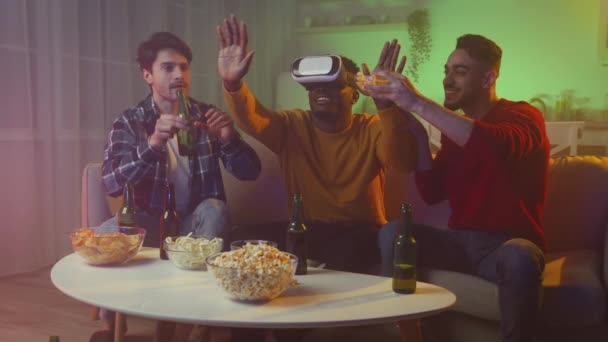 Three multiethnic men friends having fun at home in evening, one guy wearing VR glasses others joking at him, tracking shot — Video Stock