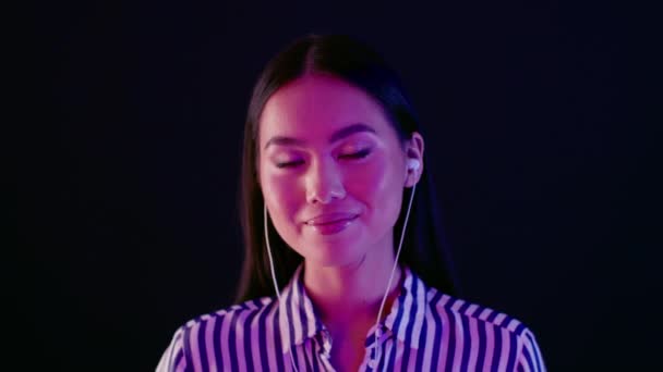 Красива Азіатська жінка слухає музику в навушниках, стоячи під яскравим неоновим світлом — стокове відео