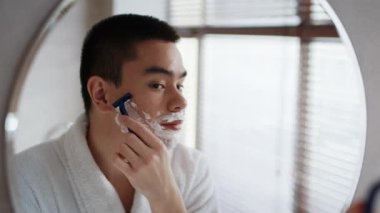 Asian Man Shaving Using Razor Standing Near Mirror In Bathroom