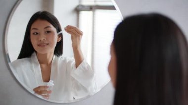 Happy Korean Lady Applying Facial Serum Standing In Bathroom