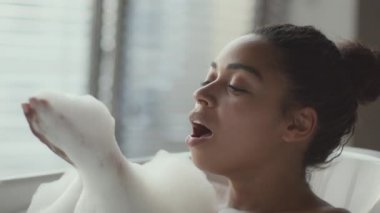Young beautiful african american woman taking bath at home, blowing foam from her palm and smiling, relaxing at bathroom