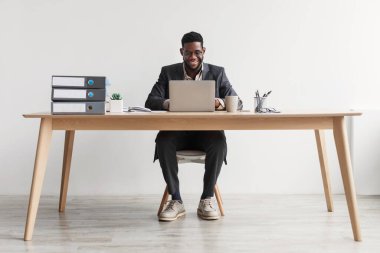 Takım elbiseli neşeli Afro-Amerikan işadamı ofiste dizüstü bilgisayarla çalışıyor.