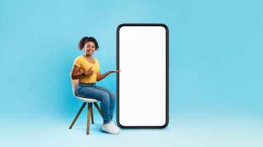 Genç Afrikalı Amerikalı kadın, mavi arka planda boş ekranlı, modern cep telefonu gösteriyor.