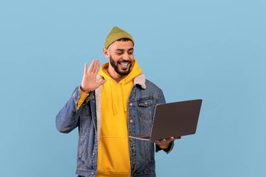 Dost canlısı Arap adam dizüstü bilgisayarla video görüşmesi yapıyor, web kamerasına el sallıyor ve gülümsüyor, mavi arka planda duruyor.