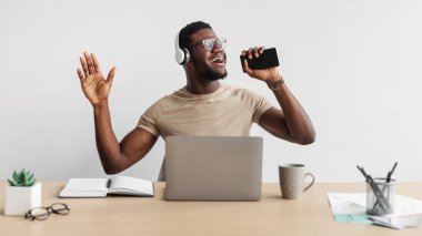 Neşeli genç siyah adam beyaz arka planda çalışırken müzik dinliyor ve akıllı telefondan şarkı söylüyor.
