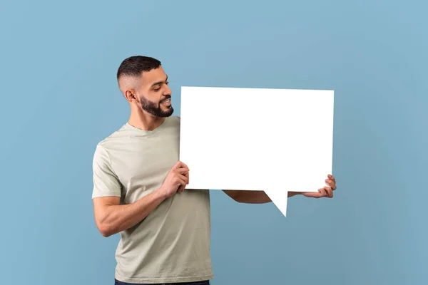 Arab man holding empty speech bubble and looking on it, standing on blue studio background, mockup — Stock fotografie