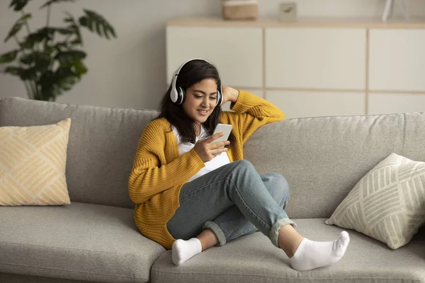 Positive arab lady in wireless headphones listening music on smartphone enjoying new online streaming app — ストック写真