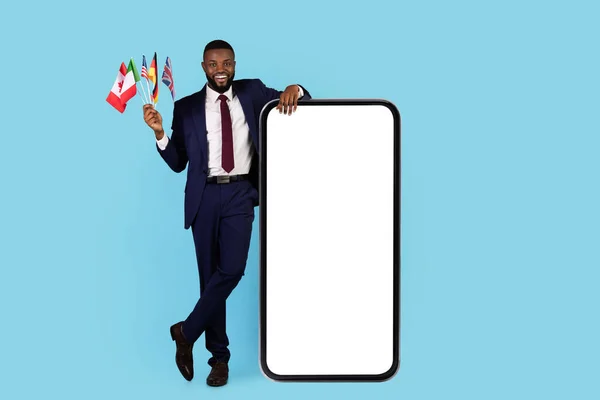 Study Languages Online. Black Businessman With Flags Standing Near Big Blank Smartphone — Stockfoto