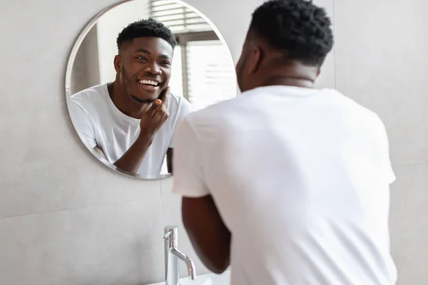 Negro chico tocando unshaven barbilla sonriendo a espejo en cuarto de baño — Foto de Stock